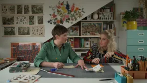 Een foto van presentator Janouk die met een kunstenaar aan tafel zit in haar atelier. Op tafel ligt er een boekje met plaatjes open en tekengereedschap. Op de achtergrond hangen er verschillende kunstwerken aan de muur.  