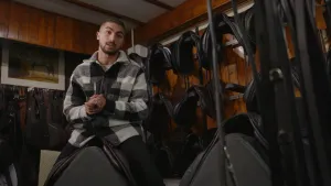 Een foto van presentator Nizar die op een zadel. Naast hem staat nog een zadel en er hangen veel zadels aan de muur op de achtergrond.