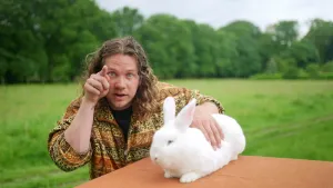 Een foto van een presentator die op een grasveld staat. De presentator wijst met zijn rechterhand naar de camera en heeft met zijn linkerhand een wit konijn vast die ligt op een tafel.