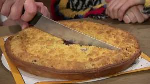 Een foto van een Limburgse vlaai die door iemand met een mes doormidden gesneden wordt.