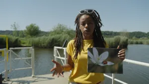 Een foto van presentator Eva die op een steiger staat van een meer. Ze houdt een foto vast van een bloem met een lieveheersbeestje erop.