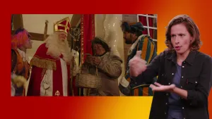 Een foto van een gebarentolk met op de achtergrond de uitzending van het Sinterklaasjournaal. In het beeld van het Sinterklaasjournaal zijn Sinterklaas en 3 Pieten zichtbaar.