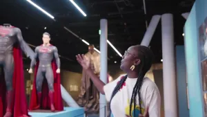 Een foto van een vrouw die met haar open hand wijst naast een groot kostuum van superman en een grote pop van superman die op een verhoging staan.