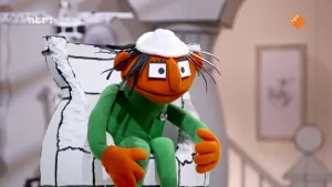 Foto van een oranje pop met groene kleding, zittend in een witte stoel.