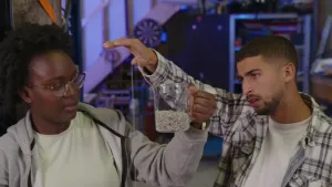 Een foto van presentatoren Tirsa en Nizar. Tirsa houdt een glazen beker vast en Nizar trekt daar een kralenketting uit. De lus van kralen die zo ontstaat heet een kralenfontein.