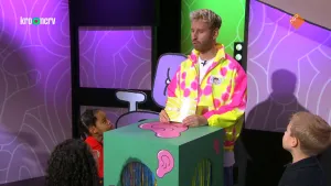 Foto van presentator in fluorescerend jack achter desk met kinderen om hem heen.