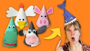 Een foto van presentator Rosalie die wijst naar haar zelfgemaakte feestmuts die lijkt op een haai. Naast haar zijn vier andere feestmutsen te zien die lijken op een kip, varken, krokodil en een koe. De achtergrond van de foto is helemaal oranje.