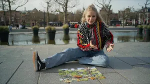 Een foto van presentator Janouk die zit op stoeptegels met een onafgemaakte puzzel. Op de puzzel zie je een groen landschap met dieren.