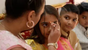 Vrouw zet met wattenstaafje een rode stip op het voorhoofd van een meisje, terwijl kinderen ernaast toekijken.