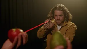 Een foto van presentator Pascal. Op de voorgrond houden twee handen een appel en een peer omhoog. Pascal richt de laserstraal van een laserpen op de appel.