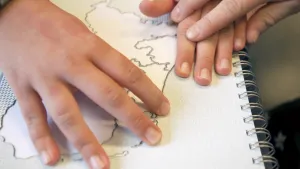 Een foto van een werkboek met een kaart van Nederland. Twee handen voelen aan de randen van de kaart. Een derde hand begeleidt de handen bij het voelen.