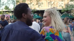 Buiten lacht Janouk naar een man. Achter hun lopen mensen en zijn parasols te zien.