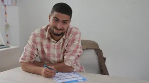 Nizar zit achter een tafel met pen en papier. Hij lacht naar de camera.