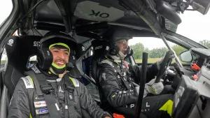 Nizar heeft een racepak aan en een helm op. Hij zit samen met nog iemand in een raceauto.