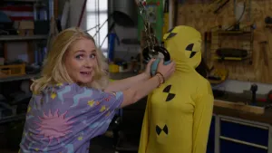 Foto van presentatrice die in de camera kijkt terwijl ze een kettlebell aan een ketting tegen het gezicht van een in het geel geklede crashtestdummie houdt.