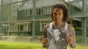 Presentatrice Astrid staat voor een groen huis dat onder een glazen kap staat. Ze heeft een zwart wit foto vast van Albert Gemmeker.