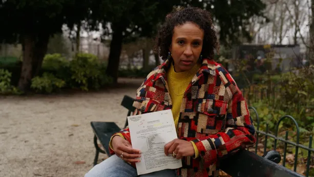 Presentatrice Astrid zit op een bankje in een park. Ze houdt een oproepbrief in haar handen.