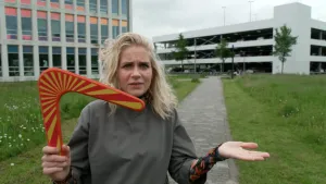 Foto van presentatrice Janouk met rood-gele boemerang in groen plantsoen voor wit gebouw.