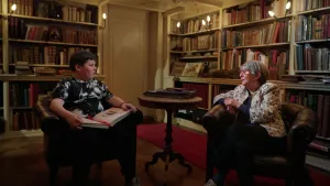Foto van 14-jarige jongen en 102-jarige vrouw in een sfeervol verlichte bibliotheek, zittend op leren stoelen. De jongen heeft een dik boek op schoot.