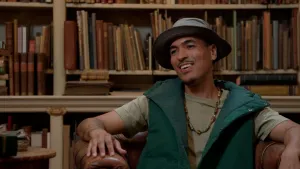 Foto van influencer Wildebras gekleed in camouflagekleuren en met een vissershoedje op, lachend op een leren stoel in bibliotheek.