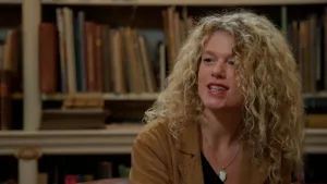 Foto van jongedame met blonde krullen, met op de achtergrond boekenkasten.