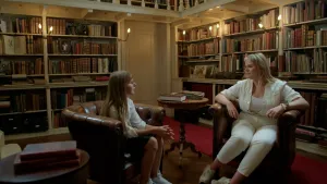 Foto van meisje in een oud uitziende bibliotheek samen met een blonde vrouw. Beide zitten op bruine leren stoelen.