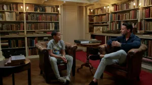 Foto van tienjarige jongen in gesprek met ongeveer 30-jarige man in een oud uitziende bibliotheek. Beide zitten op bruine leren stoelen.