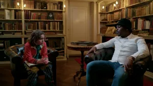 Foto van 9-jarige jongen in gesprek met een zwarte jongeman met een hoedje in een antieke bibliotheek. Beide zitten op bruine leren stoelen.