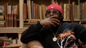 Foto van rapper Leafs, donkere man met zonnebril, rood petje en gouden ketting. Hij houdt zijn hand voor zijn mond terwijl hij nadenkt, gezeten in een bruine leren stoel met boekenkasten op de achtergrond.