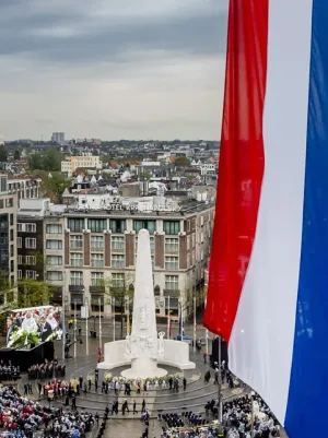 Foto van de Dam in Amsterdam, gezien vanaf het dak van het Paleis, tijdens nationale dodenherdenking. Op de voorgrond hangt de Nederlandse vlag halfstok, daarachter zie je het monument op de dam met de kransen en de toeschouwers.