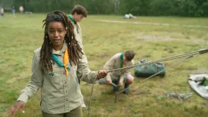 Een foto van presentator Eva in scoutingkleren. Ze staat buiten, op een grasveld, en vertelt wat in de camera. Op de achtergrond zie je twee scoutingjongens een tent opzetten.