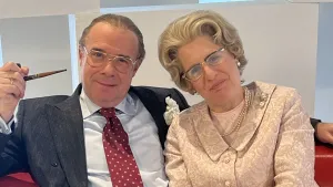 Foto van acteurs die eruit zien als prins Bernard en koningin Juliana, zittend op een bank. Bernard heeft een pijp in zijn hand.