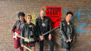 Foto van een punkband, met gitaren, drumstick, zwarte kleding, hanenkam en zwarte make-up. Op de achtergrond een muur met een poster 'Stop! Ontruiming'. en grafitti.