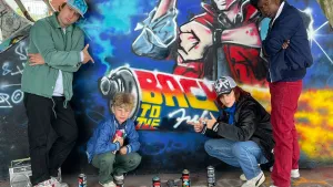 Foto van een groepje jongeren die poseert voor een muur met graffiti en op de grond allemaal spuitbussen.