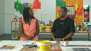 Een foto van twee mensen met een Surinaams uiterlijk. Ze zitten in een schildersatelier en zijn met elkaar in gesprek. 