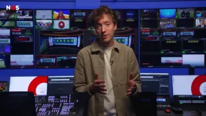 Een foto van de presentator in een regieruimte, een ruimte vol met beeldschermen. Op sommige schermen is het logo van de NOS te zien, een grote rode ronde cirkel, op andere schermen een aftelklok of een televisieprogramma.
