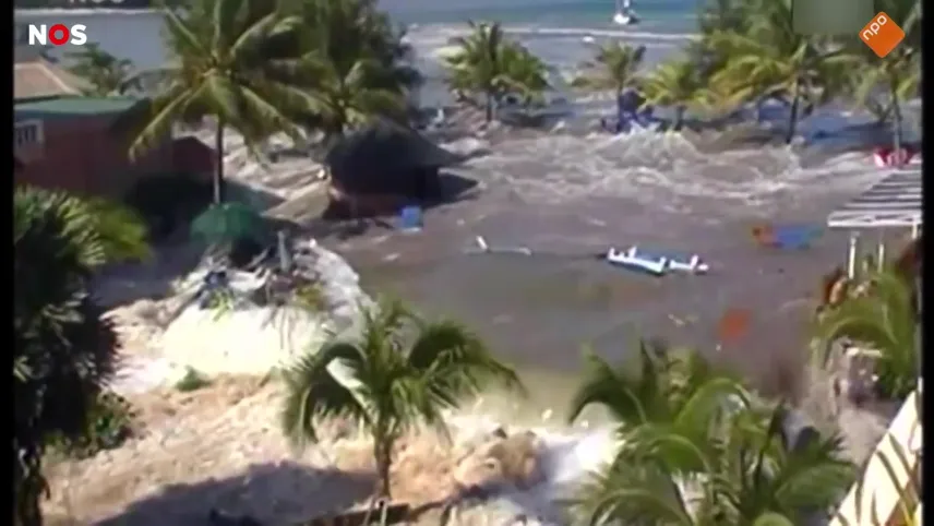 Foto van water dat een dorp overspoelt met objecten die in het water drijven. Er staan palmbomen in het water.