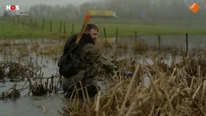 Foto van een man met stokken in een rugzak die tot zijn middel door in het water rondloopt. Op de achtergrond een dijk met auto's die eroverheen rijden.