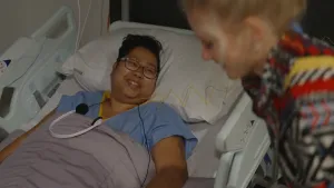 Een foto van een vrouw die in een ziekenhuisbed ligt. Er ligt een koptelefoon op haar borst. Presentatrice Janouk staat naast het bed.