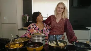 Een foto van presentator Janouk. Ze staat naast een vrouw met een Indonesisch uiterlijk in een keuken. Voor hen staan er allemaal pannen met eten erin. 