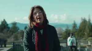 Een foto van een vrouw met een rode sjaal en een bril op, ze staat buiten met op de achtergrond naaldbomen en heuvels.