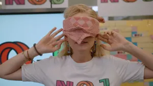 Foto van presentatrice Janouk met rode theedoek als blinddoek voor haar ogen terwijl ze haar handen naar haar oren doet om extra goed te kunnen horen.