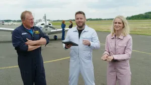 Een foto van beide presentatoren en een piloot, ze staan op een vliegveld met op de achtergrond een klein vliegtuigje, alledrie hebben ze een overall aan.