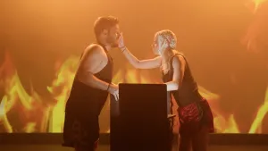 Een foto van een man en een vrouw in bokskleding, de vrouw geef de man een klap in zijn gezicht, op de achtergrond zie je een vuur geprojecteerd.