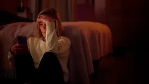 Een foto van iemand die op de grond voor haar bed zit, ze heeft een telefoon in haar hand, haar andere hand ondersteund haar voorhoofd, ze kijkt ernstig.