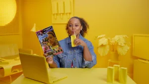 Een foto van presentatrice Quinty die een roddelblad leest en drinkt uit een geel pakje. Ook haar hele omgeving is geel, de achtergrond, laptop en andere dingen op tafel.
