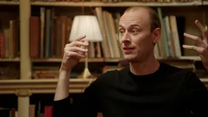 Foto van dromenexpert die met zijn handen in de lucht en gefronst gezicht iets uitlegt, in een oude bibliotheek.