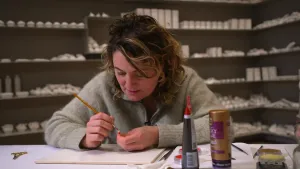 Een foto van presentator Nienke in een atelier. Ze is iets heel kleins aan het beschilderen. Ze is er met veel aandacht mee bezig. 