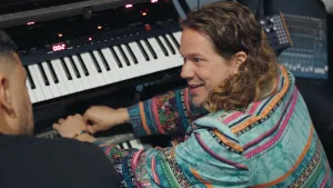 Een foto van een man achter keyboards. Hij kijkt naar een andere man die naast hem zit. 