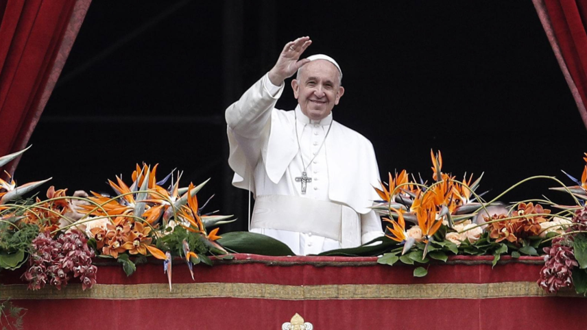 Een foto van de glimlachende en in het wit geklede paus Fransiscus die vanaf zijn versierde balkon zwaait